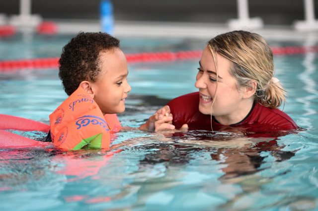 A young child in the water wearing arm bands. He is being helped by a coach in the water. They are both smiling.