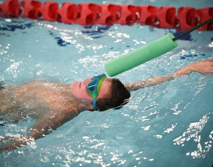 A young male swimmer taking part in the backstroke swimming race with a tapper reaching out to tap the top of his head. 